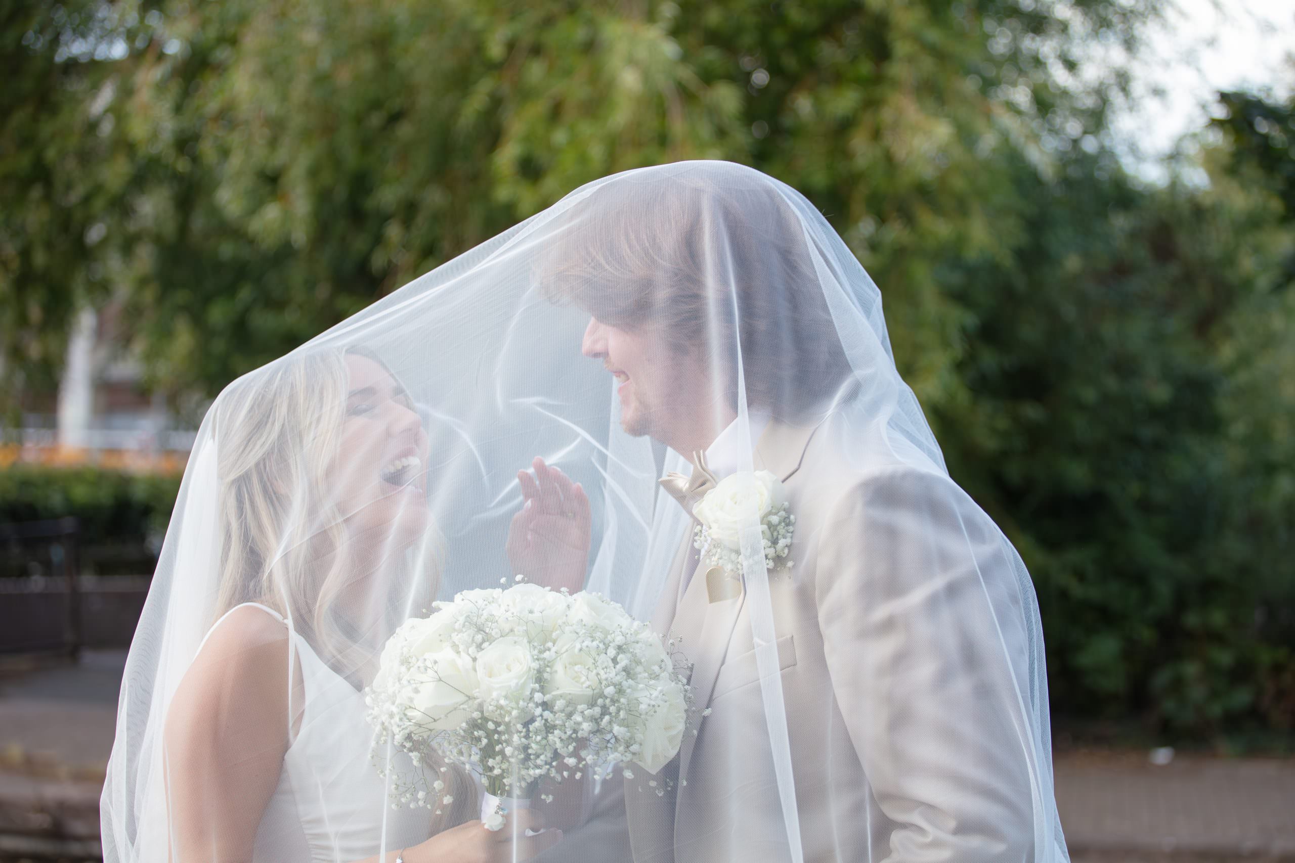 Bride and groom laugh whilst being covered by her veil.